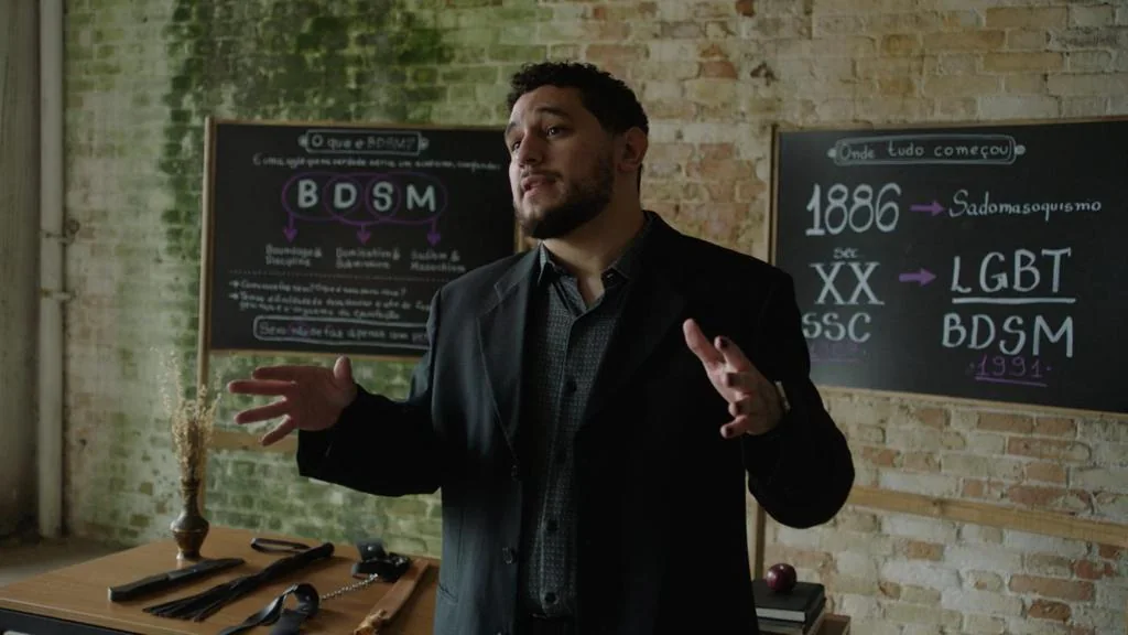 Mestre Sadic falando diante de quadros sobre a história do BDSM